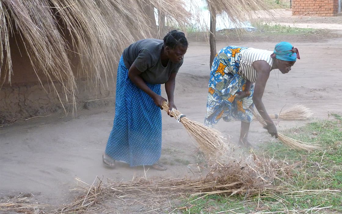 Zwei Frauen in Nabwalya fegen mit Strohbesen den staubigen Boden in der Nähe eines strohgedeckten Hauses.