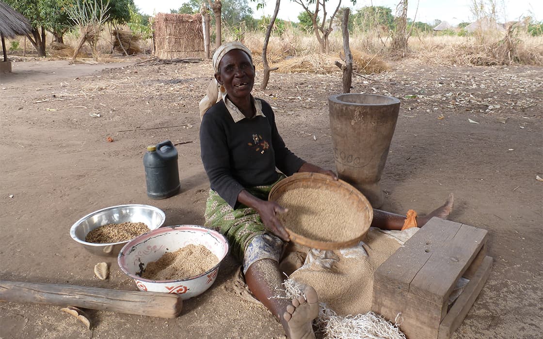 Eine Frau in Nabwalya, Sambia, lächelt, während sie mit traditionellen Werkzeugen und Schüsseln um sich herum das Getreide sortiert.