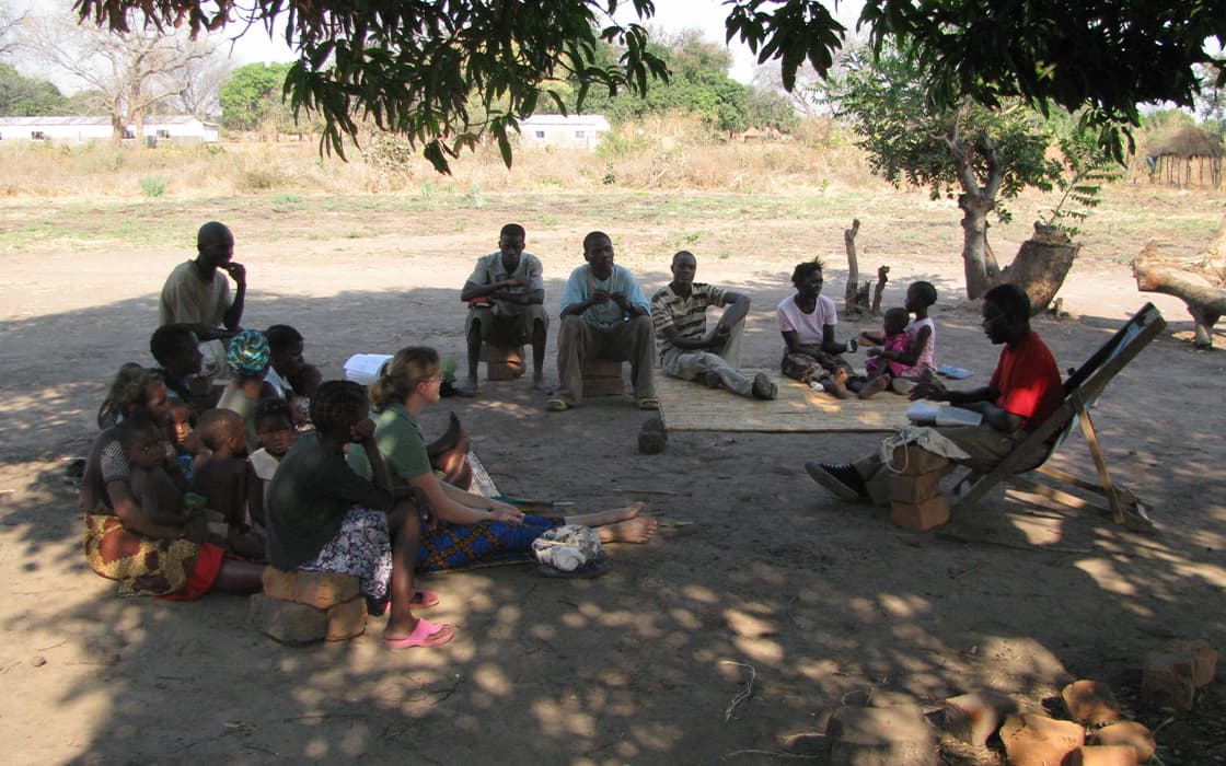 Eine Gruppe in Sambia sitzt im Freien in einem Kreis unter einem Baum, hört einem Redner zu und liest aus Papieren vor.