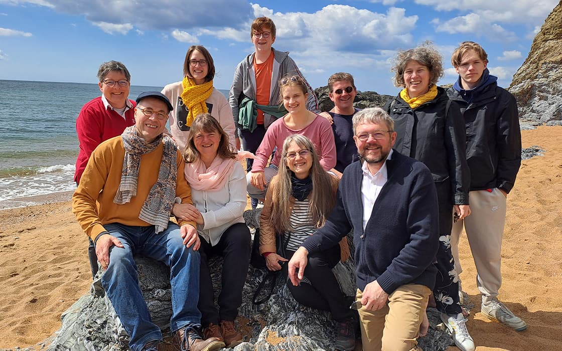 Eine Gruppe von elf Personen posiert lächelnd an einem Sandstrand in der Normandie, mit Felsen und dem Meer im Hintergrund.