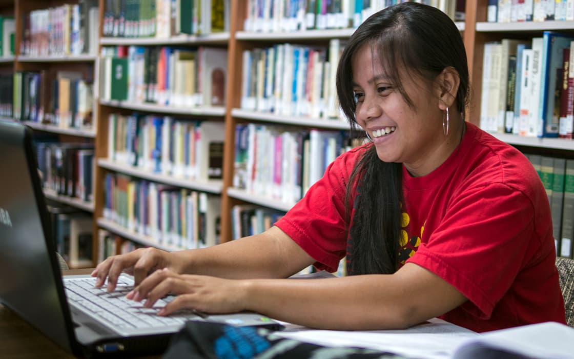Eine lächelnde Frau in einem roten Hemd tippt in der Bibliothek der Pacific Islands University auf einem Laptop, hinter ihr Bücherregale.