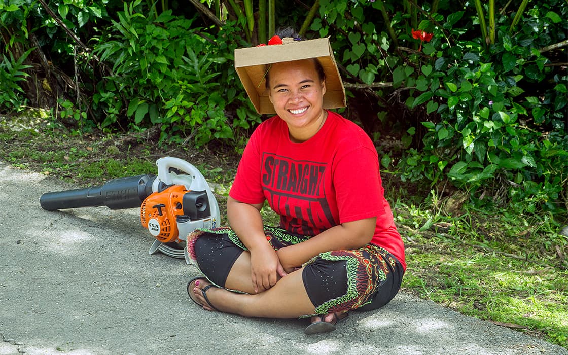 Eine lächelnde Person in einem roten Hemd und einem Hut aus Pappe sitzt auf einem Weg an der Pacific Islands University, im Hintergrund ist Grün zu sehen.
