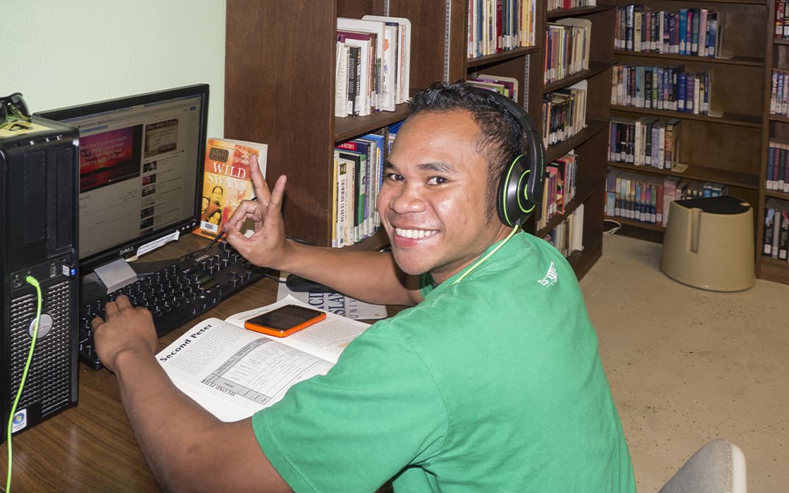 Ein lächelnder Mann mit Kopfhörern benutzt einen Computer in der Bibliothek der Pacific Islands University und hält ein Friedenszeichen hoch.