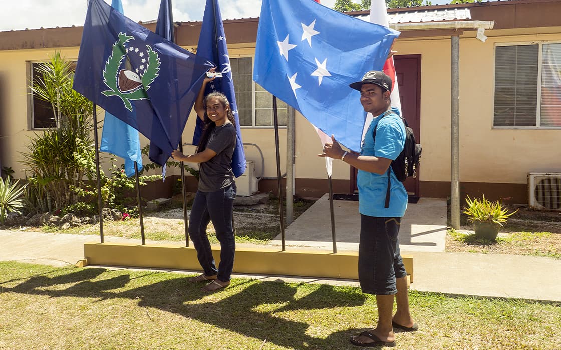 Zwei Personen stehen an einem sonnigen Tag in Mikronesien vor der Pacific Islands University und halten blaue Fahnen in die Kamera.