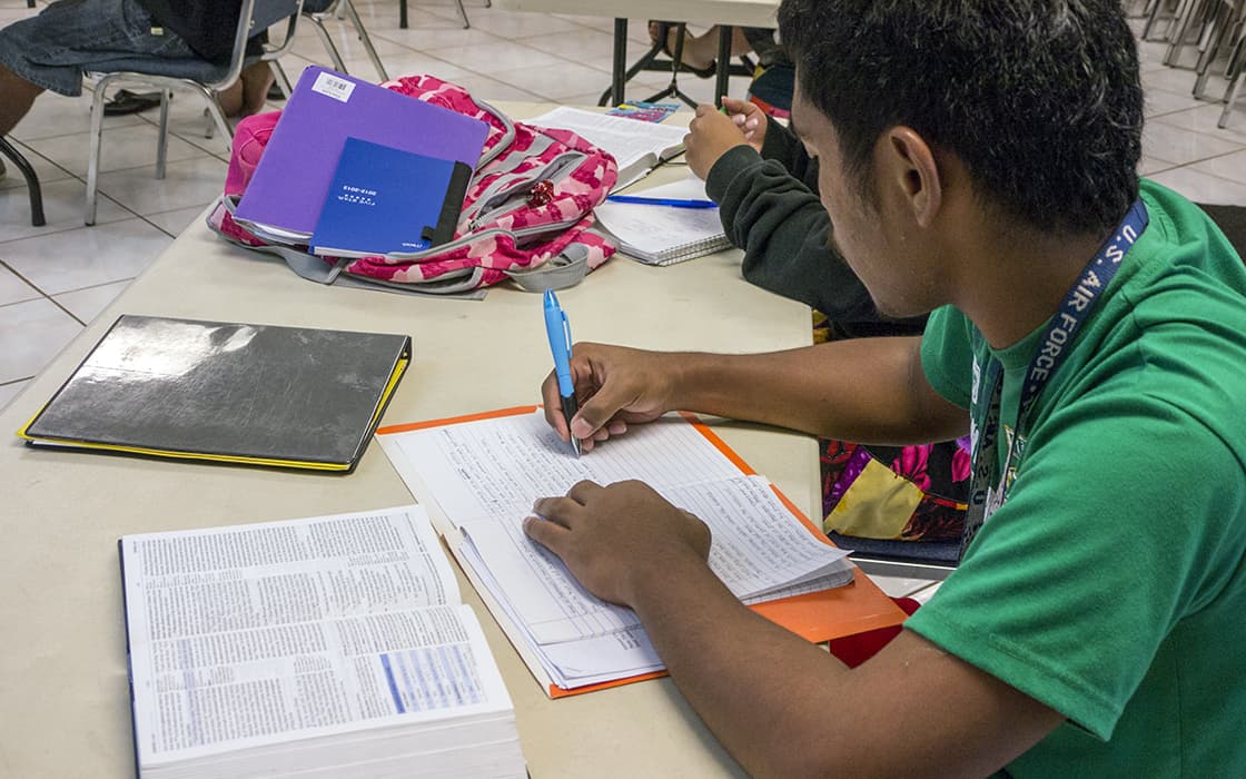 Ein Student der Pacific Islands University schreibt in ein Notizbuch an einem Tisch, an dem Bücher, ein Ordner und ein rosa Rucksack liegen.
