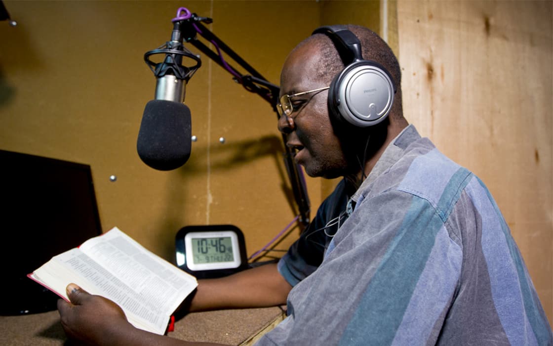 Ein Mann mit Kopfhörern liest aus einem Buch in ein Mikrofon im Studio von Radio L in Malawi. Eine Uhr zeigt 10:46.