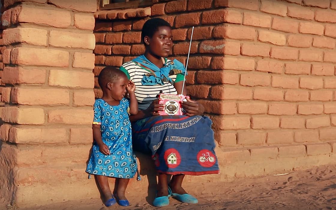 Eine Frau und ein Kind in Blau sitzen an einer Backsteinmauer, während sie in ihrem Radio Radio L Malawi hören.