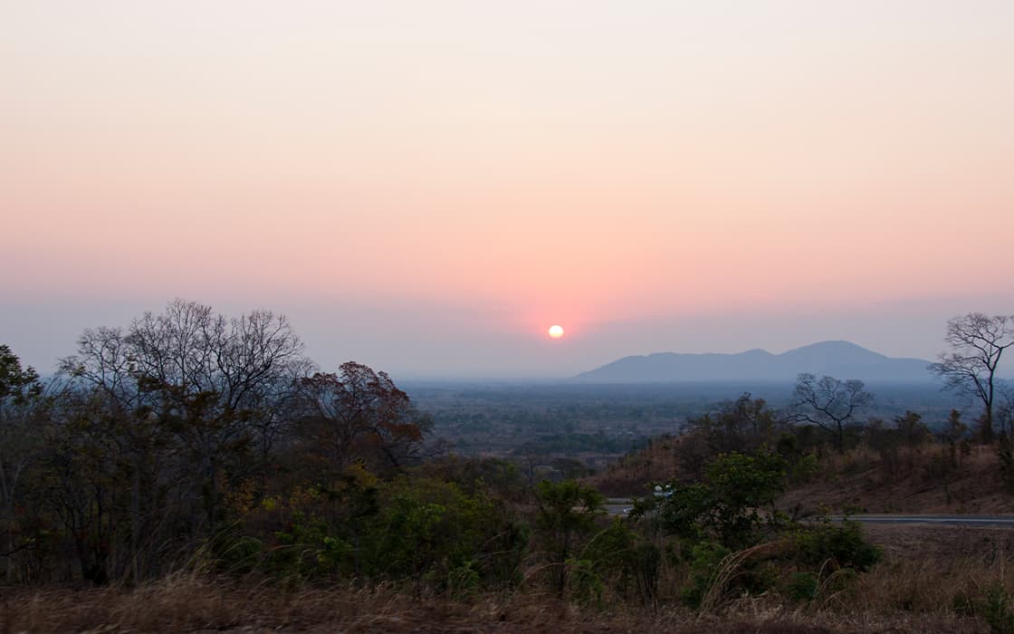 Sonnenuntergang über einer trockenen Landschaft mit spärlichen Bäumen und fernen Hügeln unter einem dunstigen Himmel, der die ruhige Stimmung von Radio L Malawi widerspiegelt.