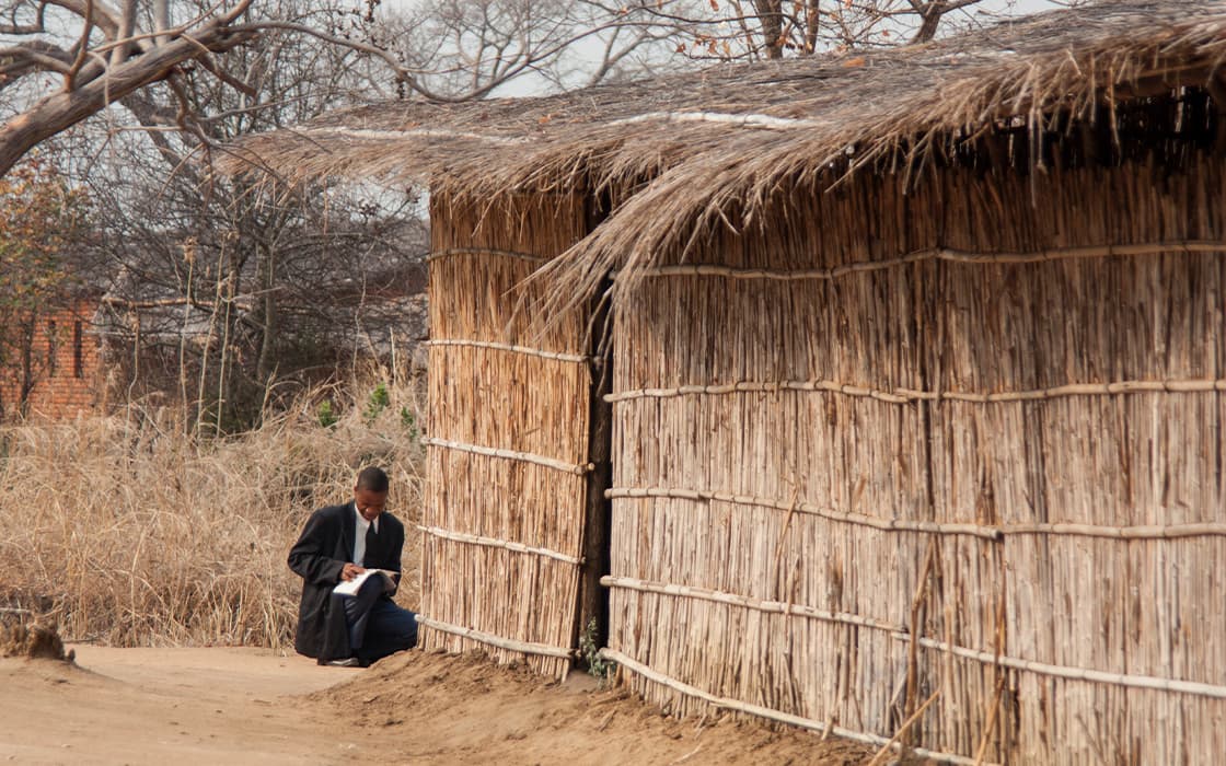 Eine Person sitzt mit einem Buch neben einer strohgedeckten Hütte aus Stöcken und Stroh im ländlichen Malawi.