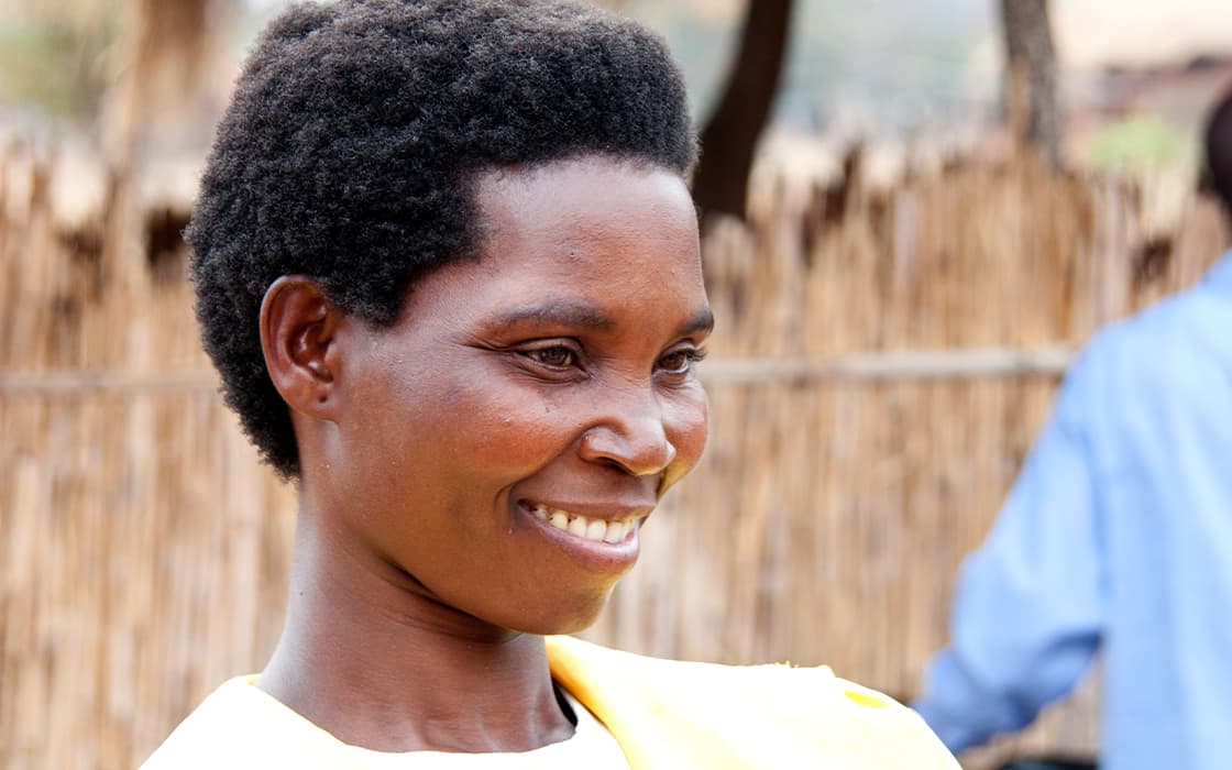 Eine Frau mit kurzen lockigen Haaren lächelt im Freien vor einem Bambuszaun in Malawi freundlich.