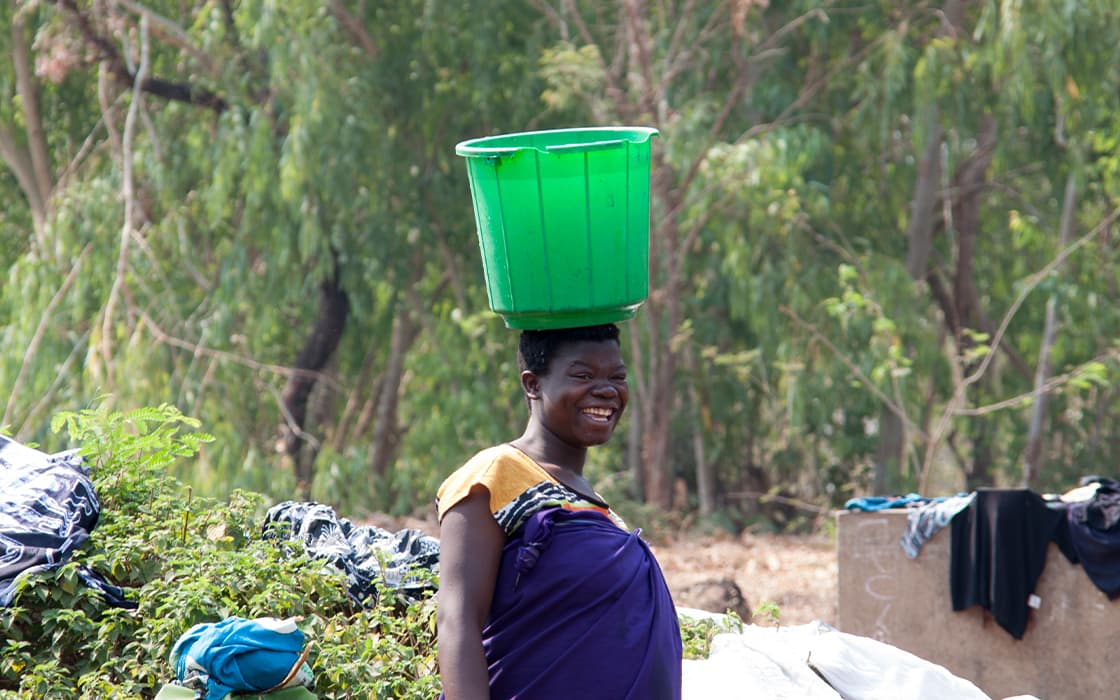 Eine lächelnde Frau in Malawi trägt einen großen grünen Eimer auf dem Kopf, umgeben von Grünzeug und Wäsche.