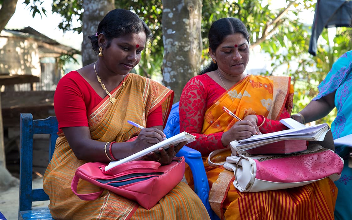 Zwei Frauen in bunten Saris, die zu TEE Bangladesch gehören, sitzen im Freien und schreiben in Notizbücher für die theologische Ausbildung.