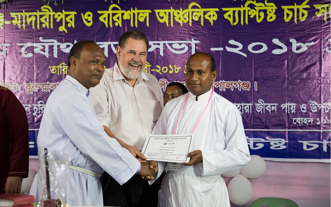 Drei Männer stehen zusammen in Bangladesch, zwei in weißen Gewändern und schütteln sich die Hände bei einer TEE-Zeremonie zur theologischen Ausbildung.