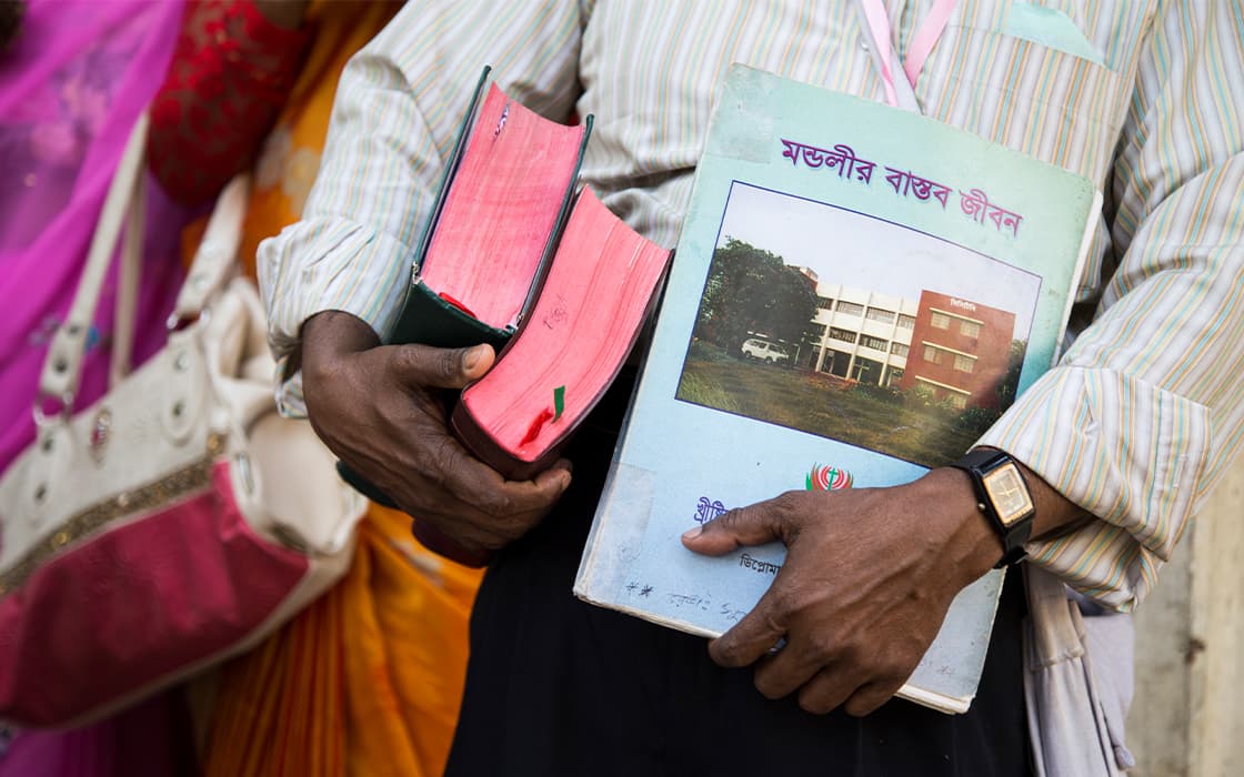 Eine Person hält zwei dicke rote Bücher und einen blauen Ordner über TEE Bangladesch mit Text in Assamesisch auf dem Umschlag.
