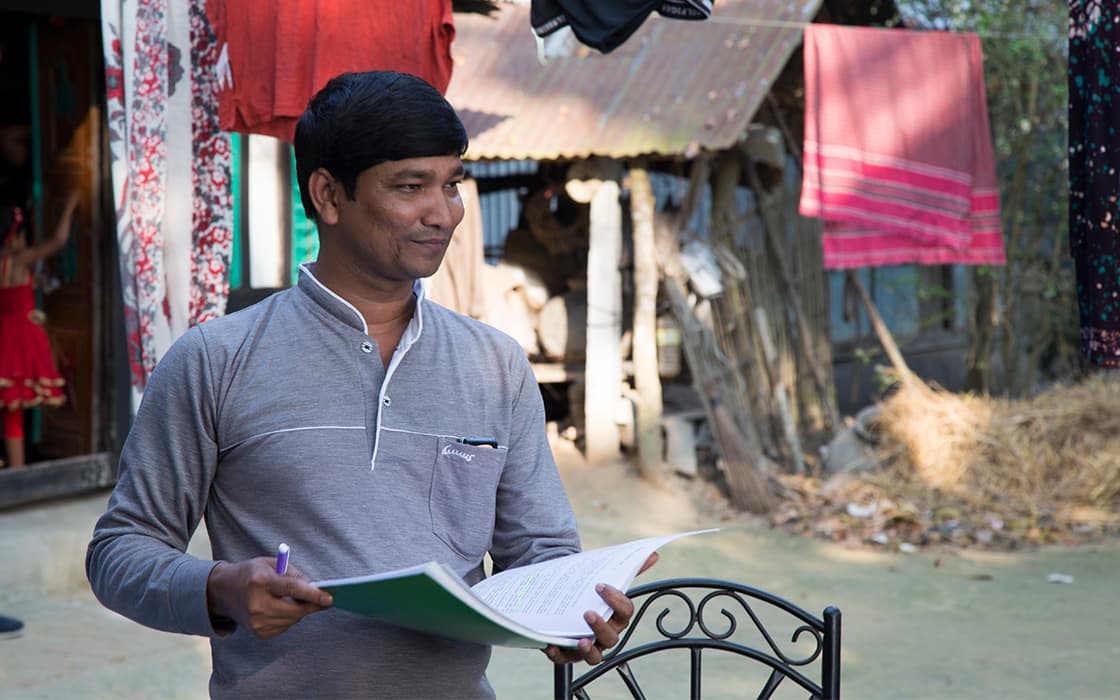Ein Mann steht im Freien mit Papieren und einem Stift, inmitten von Wäsche und rustikalen Gebäuden, die TEE Bangladesch symbolisieren.