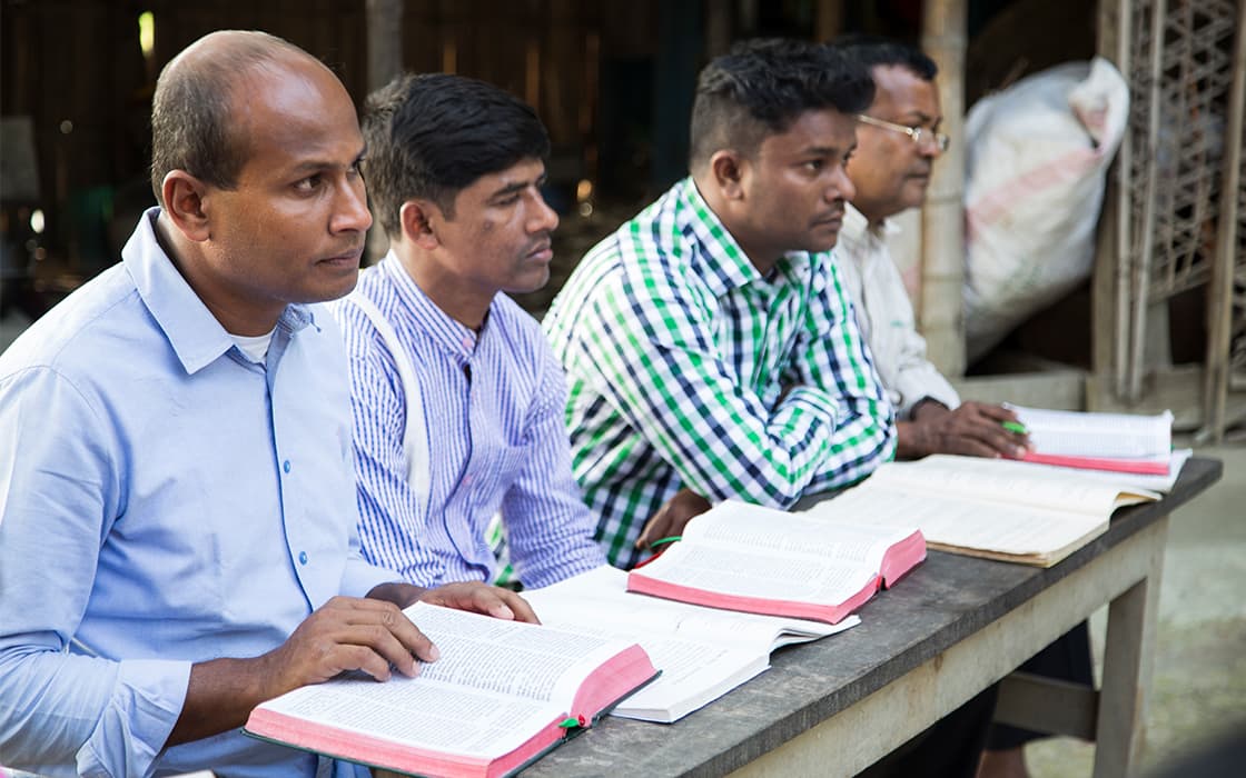 Vier Männer in Bangladesch sitzen im Freien an einem Tisch, konzentriert auf ihre theologische Ausbildung mit offenen Büchern.
