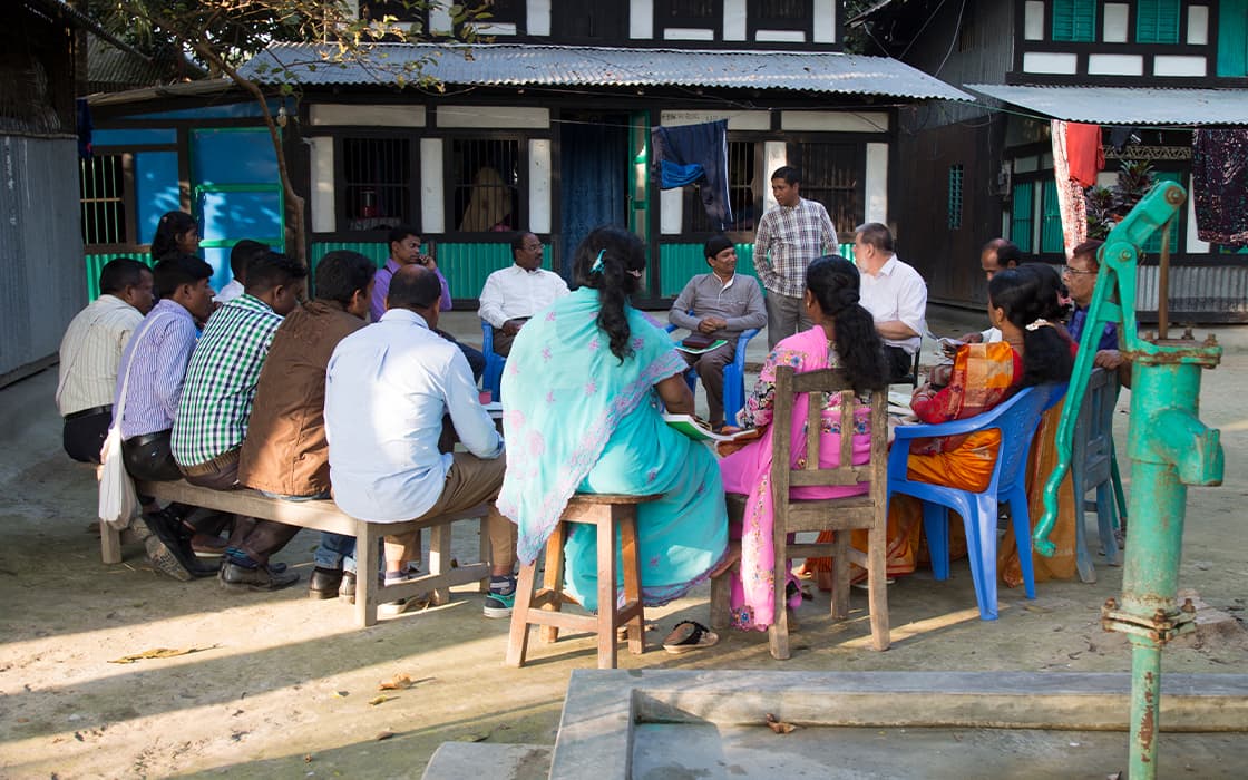 Eine Gruppe diskutiert über außerschulische theologische Ausbildung in der Nähe eines Gebäudes und einer Wasserpumpe in TEE Bangladesch.