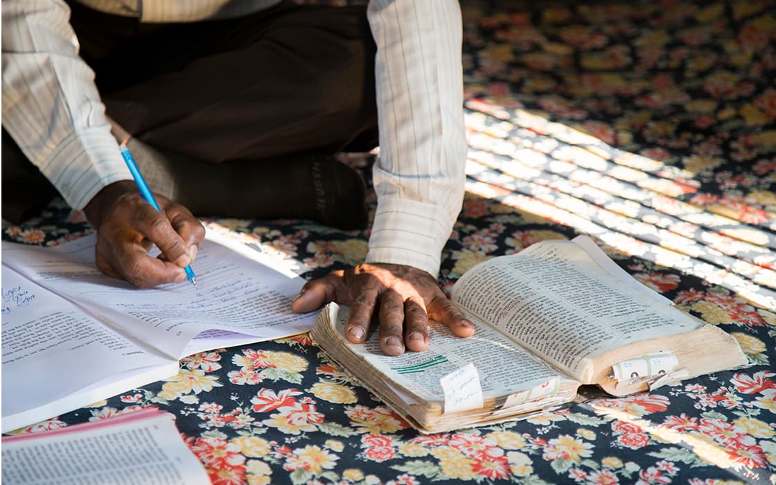 Eine Person studiert für eine theologische Ausbildung und schreibt in ein Notizbuch mit TEE-Materialien auf einem blumengemusterten Stoff.