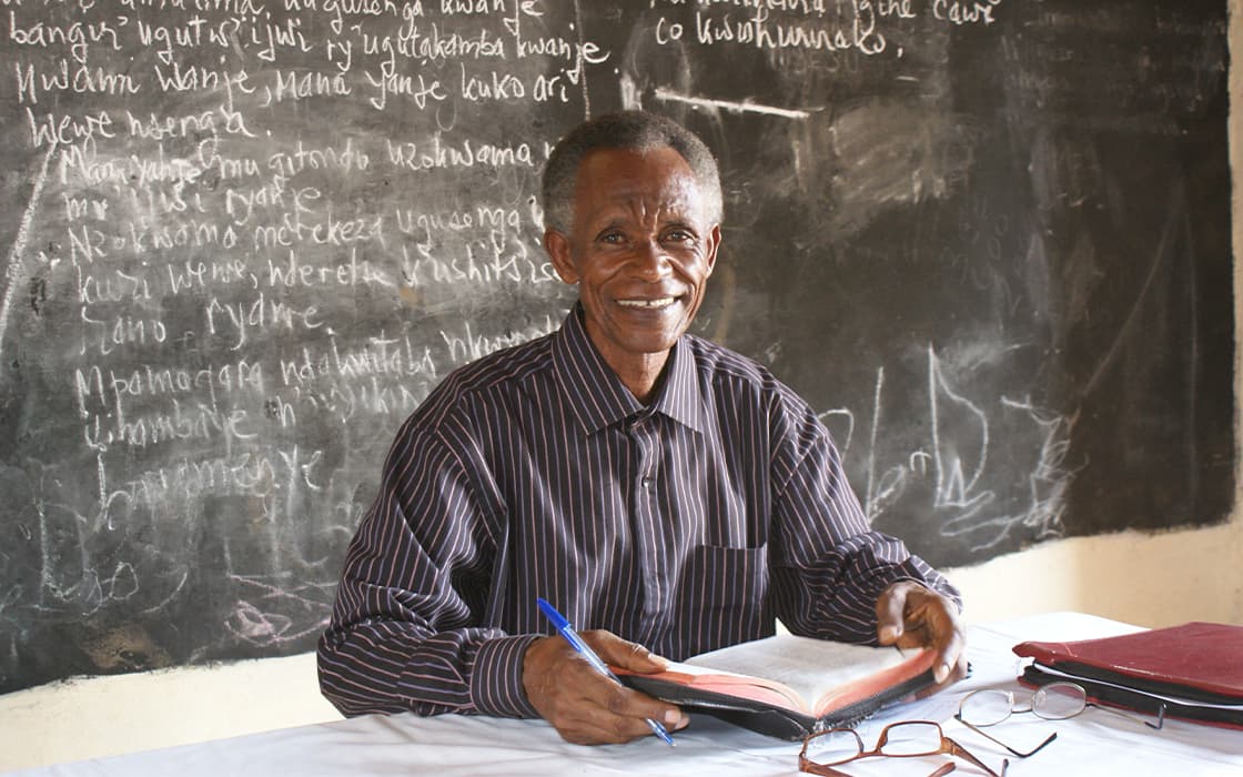 Lächelnder Mann im gestreiften Hemd studiert für TEE Burundi theologische Ausbildung an einem Schreibtisch, Kreidetafel hinter ihm.