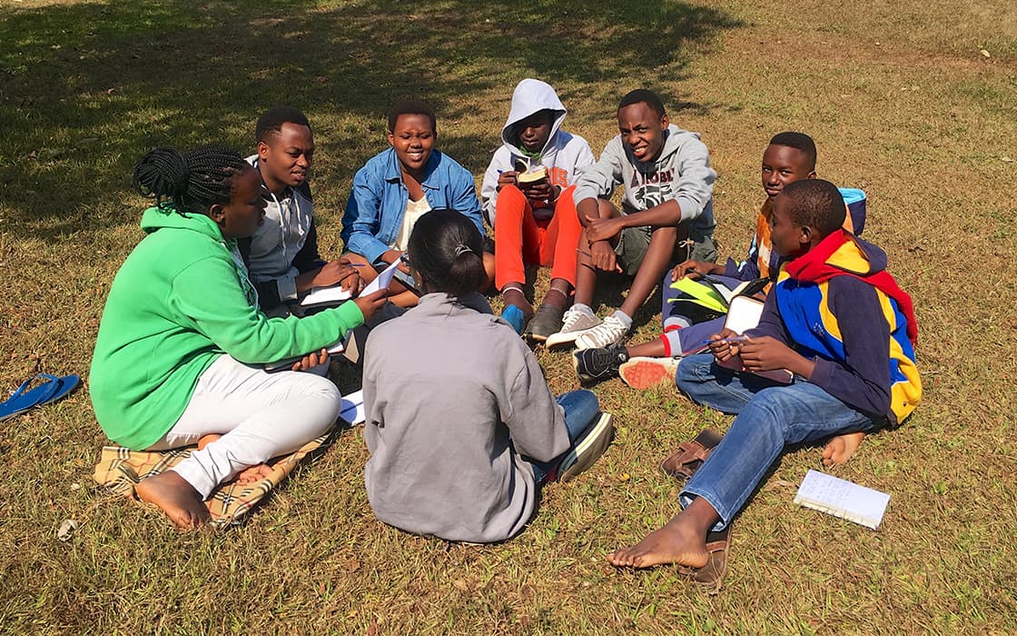 Acht junge Leute sitzen im Kreis auf einer Wiese, diskutieren über die theologische Ausbildung und schreiben Notizen an einem sonnigen außerschulischen Tag.