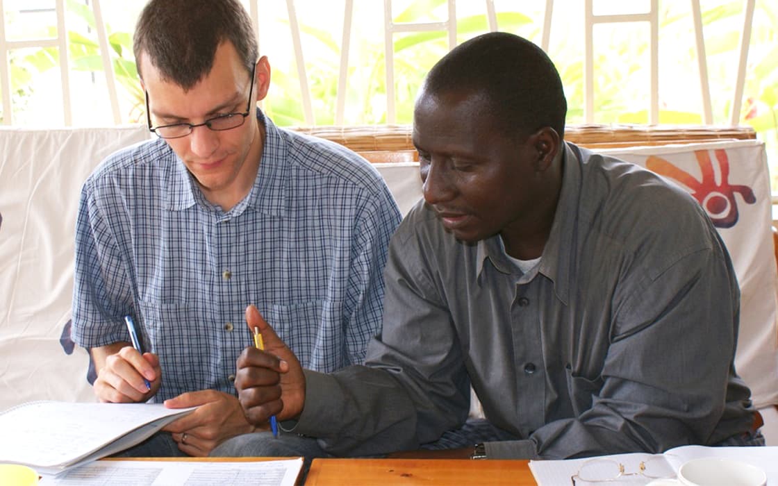 Zwei Männer diskutieren Dokumente und machen sich Notizen zur theologischen Ausbildung am TEE Burundi in einem hellen Innenraum.