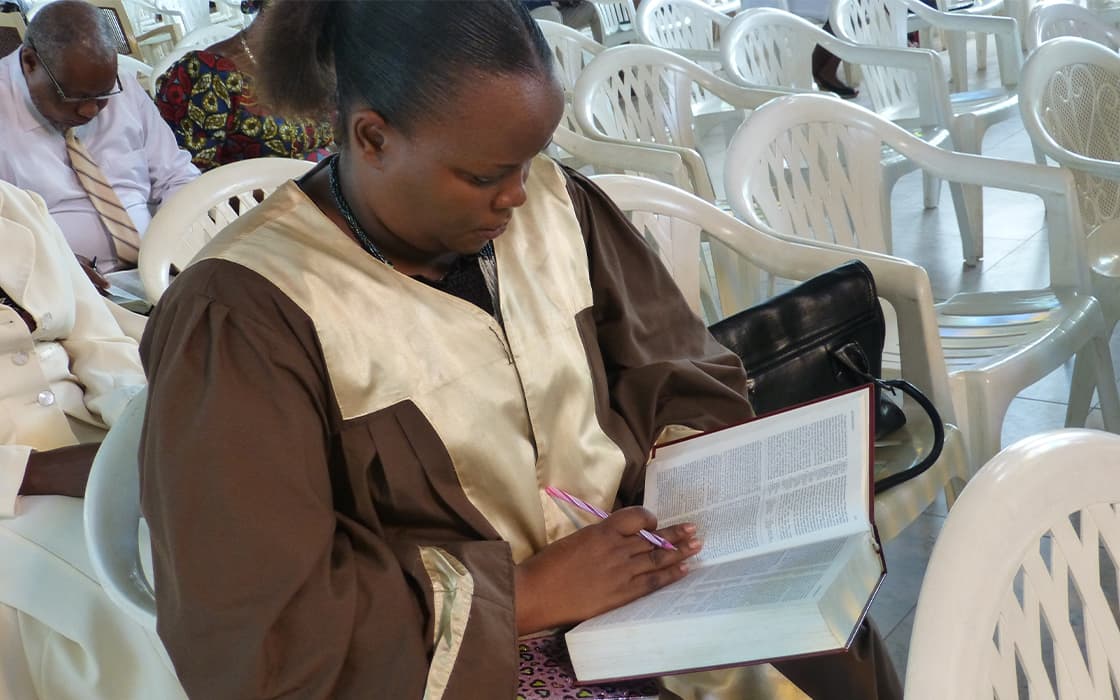 Eine Frau im Bademantel schreibt in einem Buch bei einer Versammlung des TEE Burundi zur theologischen Ausbildung inmitten von weißen Plastikstühlen.