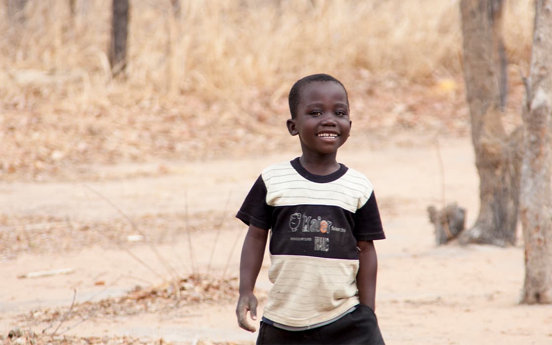 Ein kleiner Junge lächelt, während er in Malawi im Freien steht, umgeben von trockenem Gras und Bäumen in einer ländlichen Umgebung.
