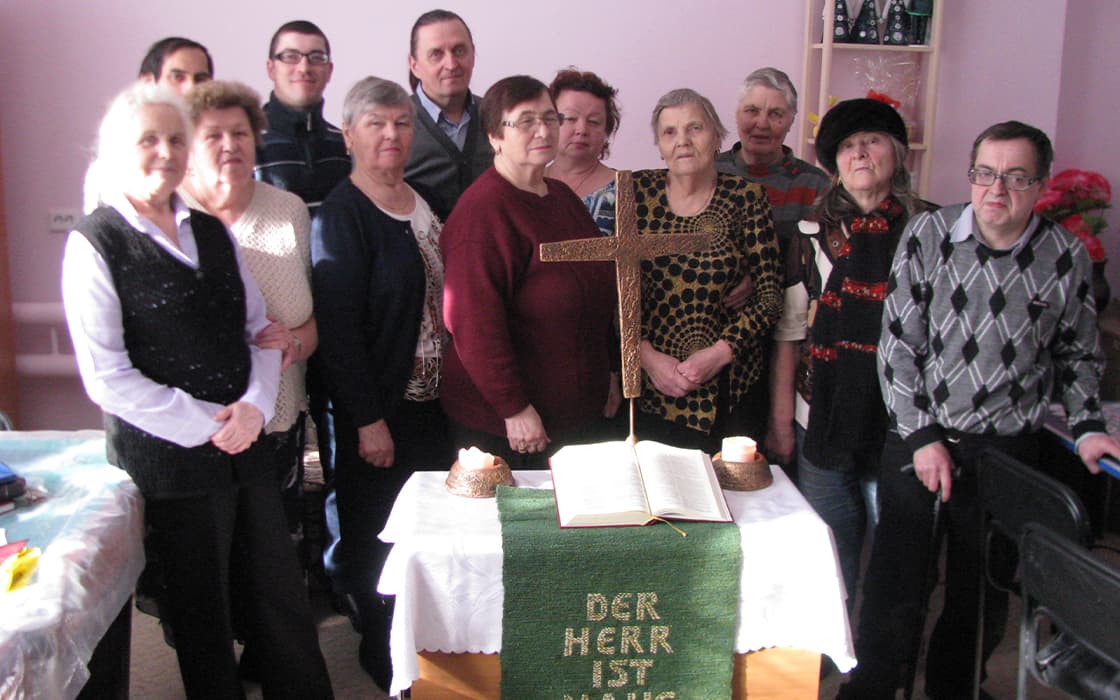 Eine Gruppe von zwölf Personen steht hinter einem Tisch mit einer aufgeschlagenen Bibel und Symbolen der theologischen Ausbildung.