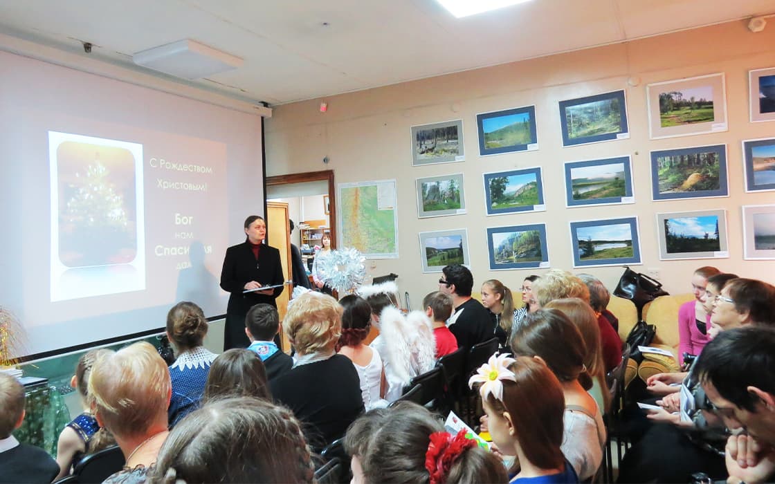 Ein Mann hält vor einem sitzenden Publikum in einem Klassenzimmer einen Vortrag über die theologische Ausbildung in Russland, wobei ein Weihnachtsdia projiziert wird.