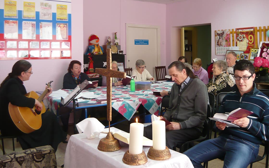 Eine kleine Gruppe in Russland sitzt bei Kerzenlicht, singt und liest aus Büchern; eine Person spielt Gitarre.