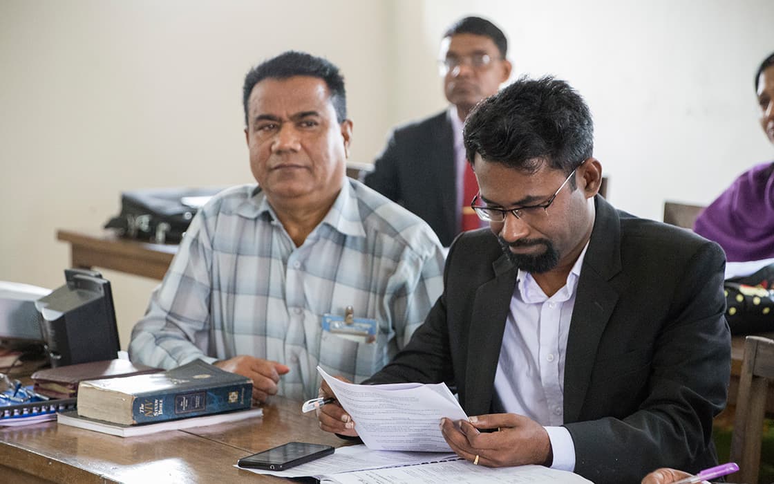 Zwei Männer sitzen an einem Schreibtisch mit Papieren; der eine liest, während der andere aufschaut, er studiert am College of Christian Theology Bangladesh.