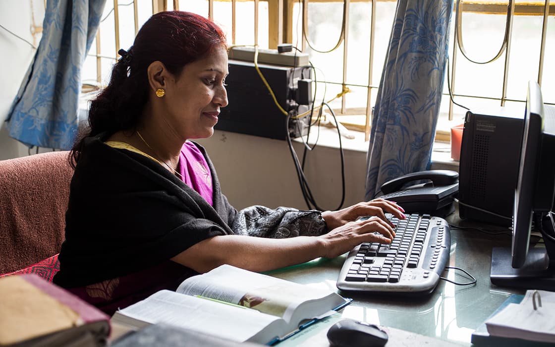 Eine Frau tippt an einem Schreibtisch inmitten von Büchern und Papieren und studiert in einem hellen Raum in Bangladesch christliche Erziehung.