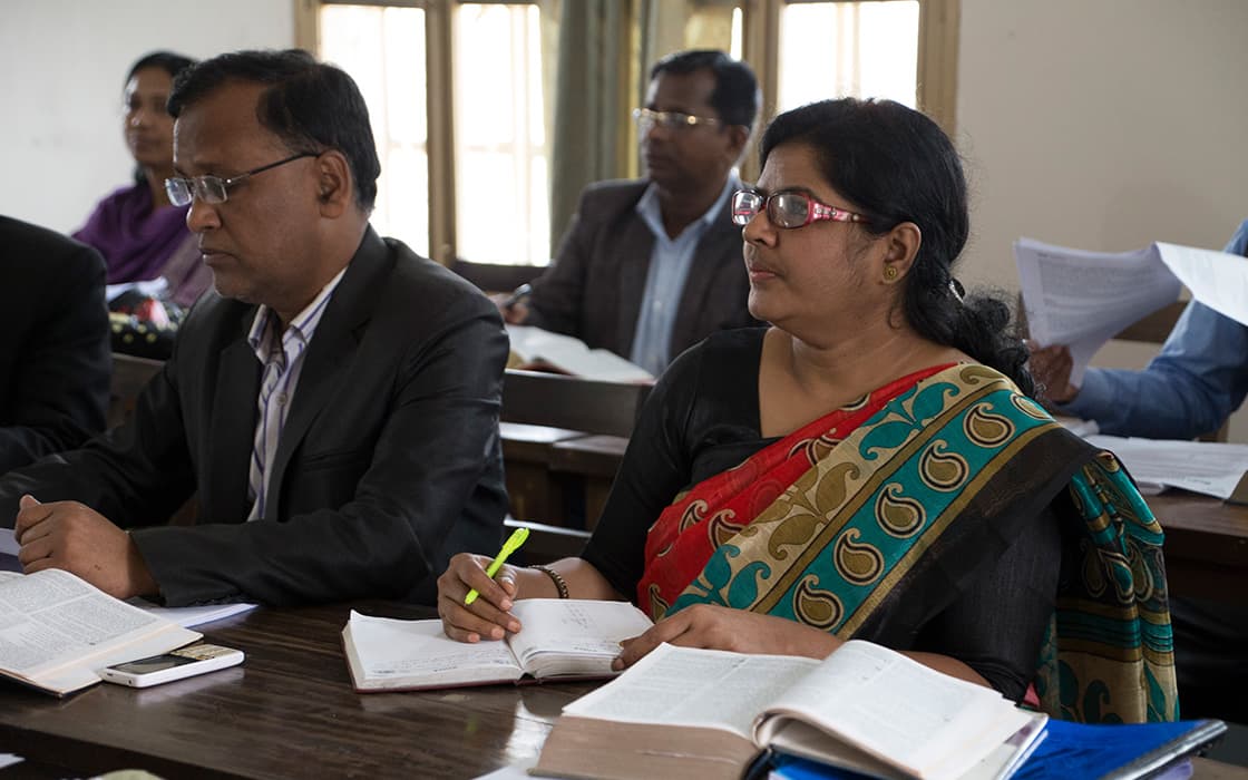 Zwei Erwachsene sitzen in einem Klassenzimmer der Hochschule für Christliche Theologie in Bangladesch an den Tischen und machen sich während einer Vorlesung Notizen.