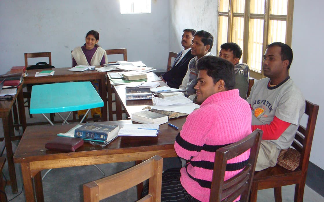 Sechs Erwachsene sitzen in einem Klassenzimmer der Christlichen Theologie Bangladeschs und haben Bücher und Papiere vor sich ausgebreitet.