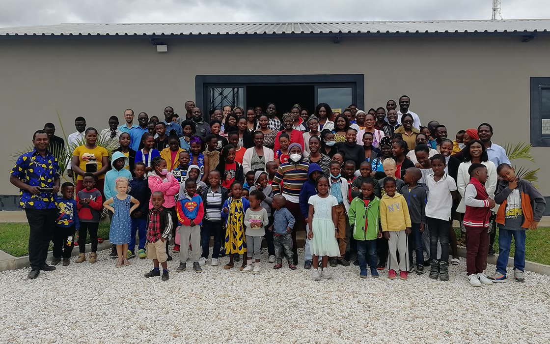Eine große Gruppe von Erwachsenen und Kindern in Sambia posiert vor einem Gebäude und lächelt an einem bewölkten Tag in die Kamera.