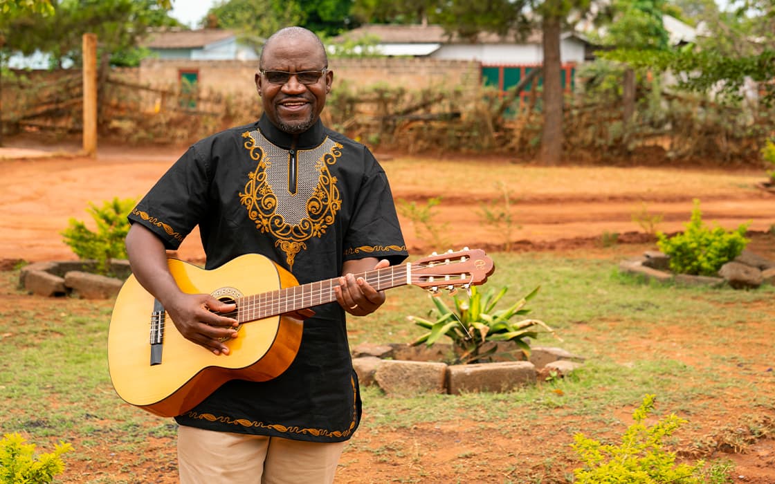 Mann im schwarz-goldenen Hemd spielt Akustikgitarre im Freien in Sambia, lächelnd, mit Gemeindegründungen im Hintergrund.