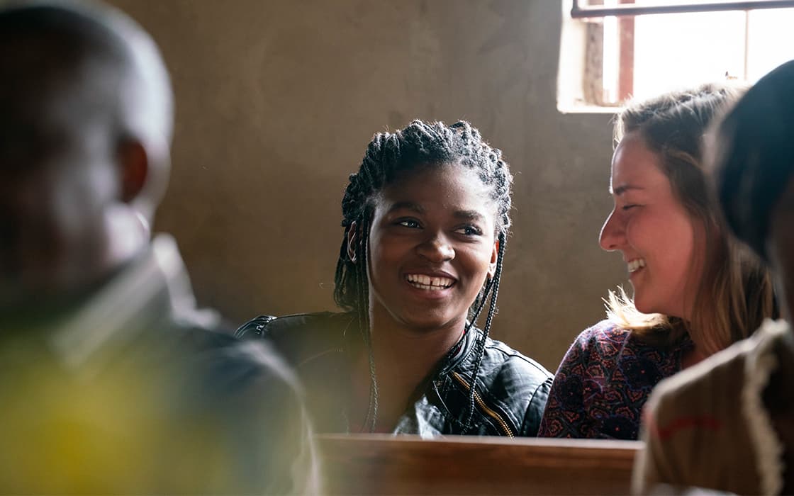 Zwei Frauen sitzen zusammen auf einer Bank in Sambia und lächeln und lachen, während das Sonnenlicht durch das Fenster hinter ihnen fällt.