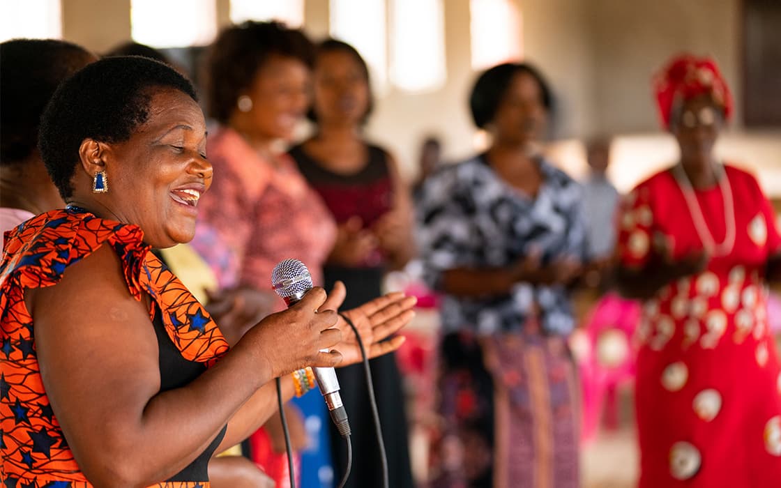 Eine lächelnde Frau singt in Sambia in ein Mikrofon, während im Hintergrund mehrere Frauen stehen und klatschen.