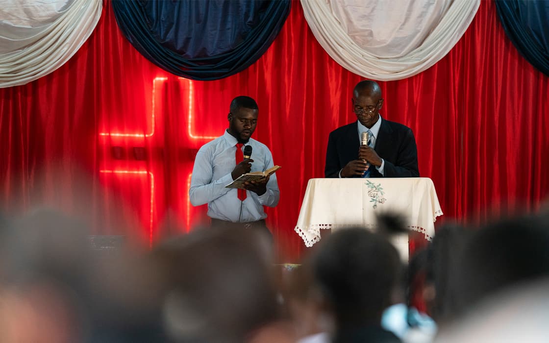 Zwei Männer sprechen an einem Kirchenpodium in Sambia; einer liest aus einem Buch. Ein roter Hintergrund und ein Kreuz heben Gemeindegründungen hervor.