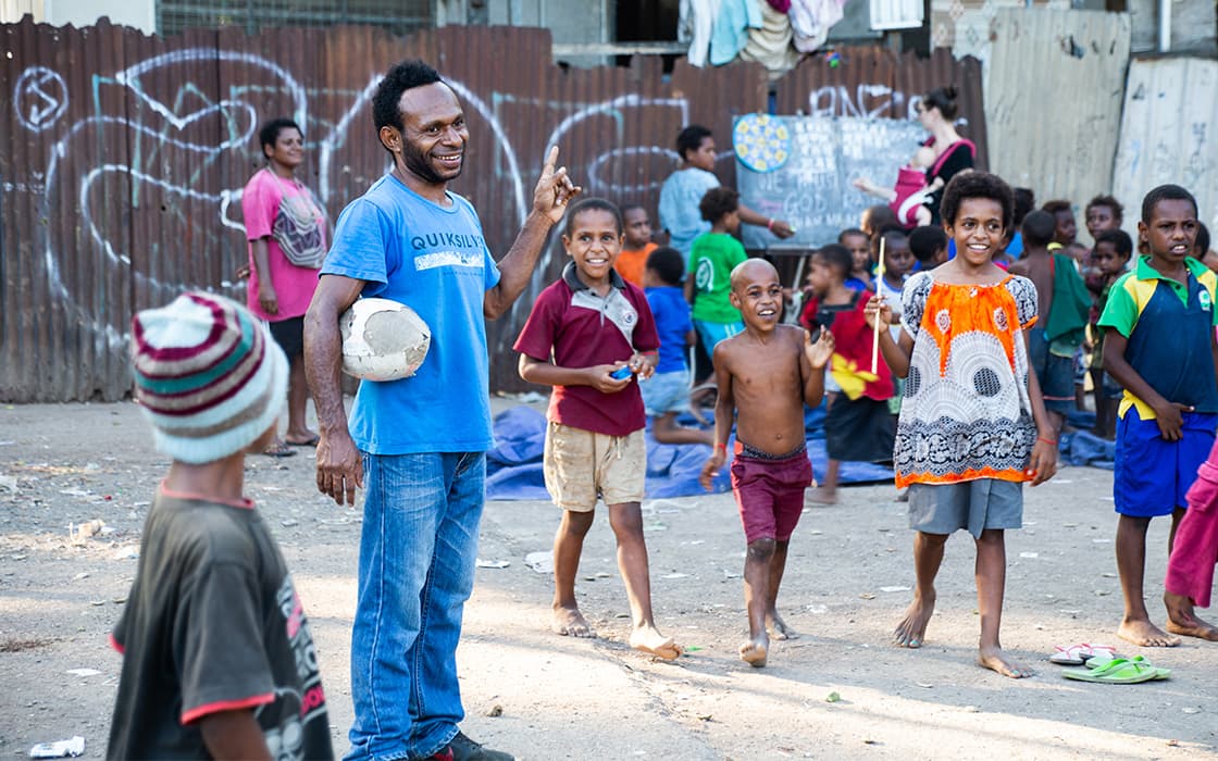 Eine Gruppe lächelnder Kinder und ein Mann mit einem Fußball stehen zusammen im Freien in Papua-Neuguinea.