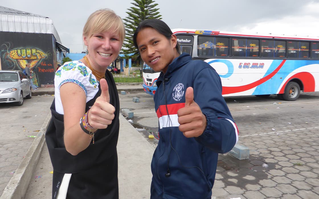 Zwei Personen lächeln und zeigen die Daumen nach oben in der Nähe eines Busses und genießen ihre Erfahrung mit Studentenarbeit in Ecuador.