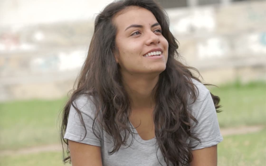 Eine junge Frau mit langen braunen Haaren und grauem Hemd lächelt, während sie die Jugendarbeit Ecuador im Freien auf einer Wiese genießt.