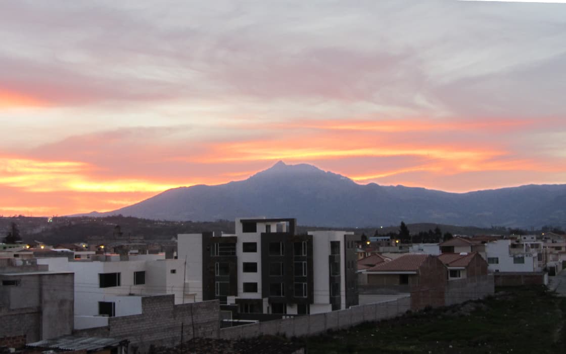 Sonnenuntergangshimmel mit orangefarbenen Wolken über einem Berg, Wohngebäude im Vordergrund - Studentenarbeit Ecuador.