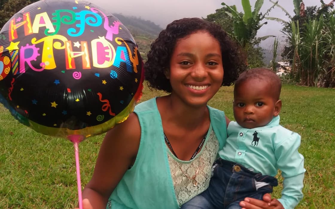 Eine lächelnde Frau hält einen Geburtstagsluftballon und ein Baby in der Hand, während sie im Freien im Casa Ágape Ecuador im Gras sitzt.