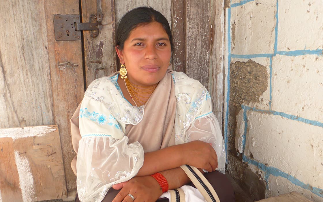 Eine Frau in traditioneller Kleidung und Goldschmuck sitzt an einer rustikalen Tür im Casa Ágape Ecuador und schaut in die Kamera.