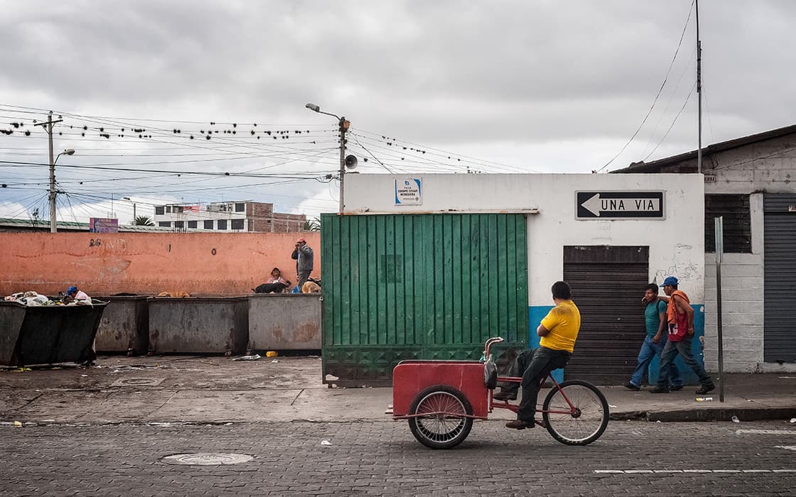 Mann in gelbem Hemd auf rotem Dreirad in der Nähe des Gebäudes; Menschen fahren an Mülleimern vorbei, Casa Ágape Ecuador unterstützt Hilfe für Frauen.