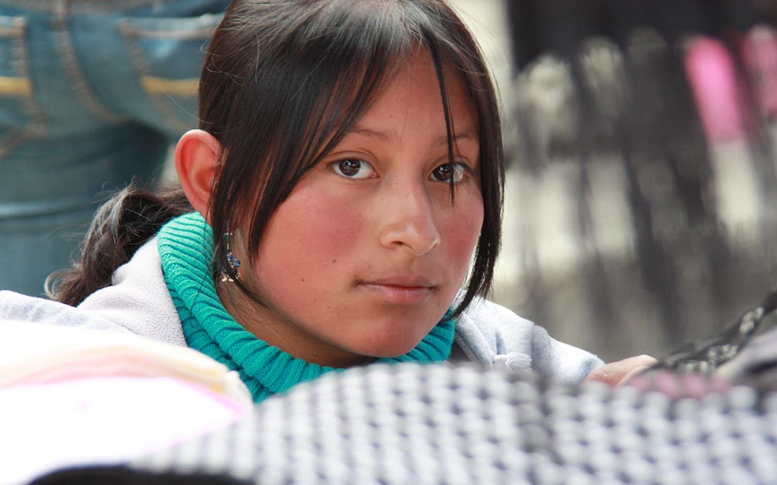 Junge Frau mit dunklem Haar und rosigen Wangen, trägt einen türkisfarbenen Pullover und eine graue Jacke - mit Unterstützung von Casa Ágape Ecuador.