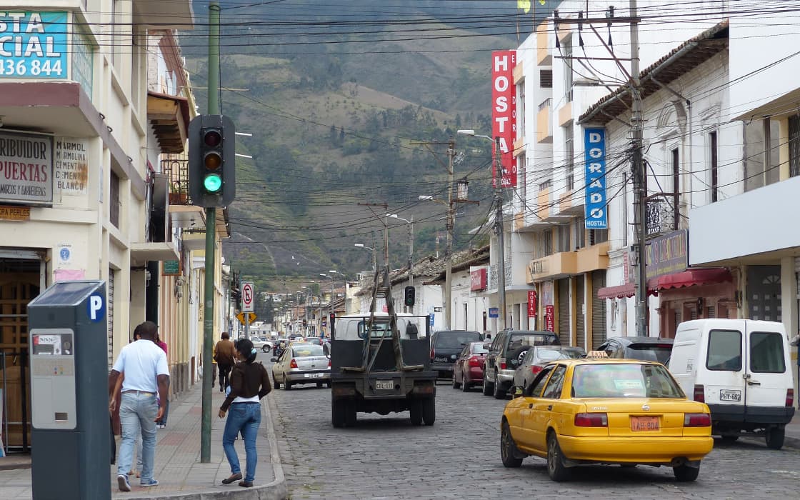 Eine Straßenszene in einer kleinen Stadt in der Nähe von Casa Ágape Ecuador, mit Autos, Fußgängern, Geschäften und Bergen im Hintergrund.