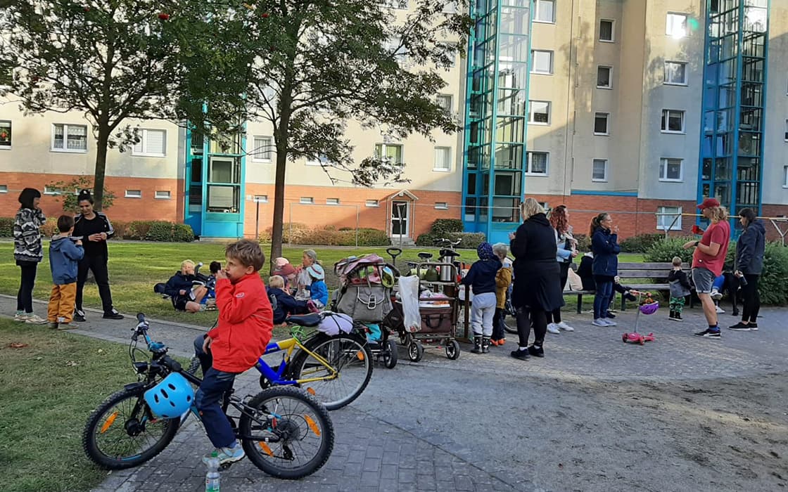Kinder und Erwachsene versammeln sich im Freien in der Nähe eines Wohnhauses, mit Fahrrädern, Kinderwagen und Bäumen in der Nähe.