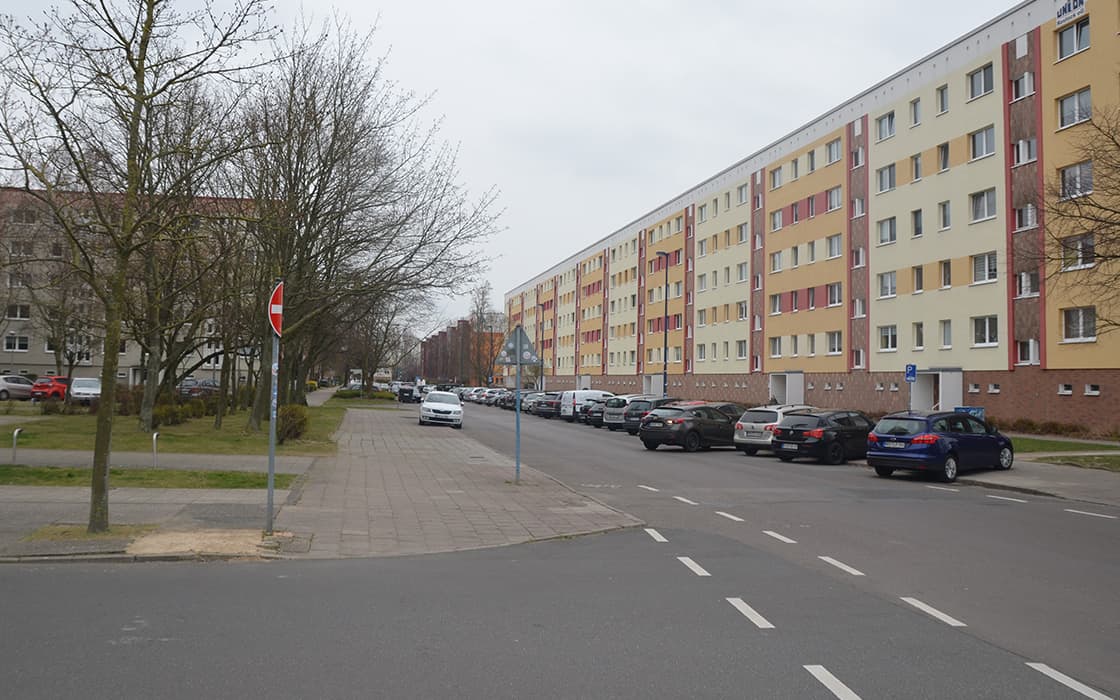 Eine Straße in Rostock mit geparkten Autos säumt an einem bedeckten Tag ein langes, mehrstöckiges Wohnhaus.